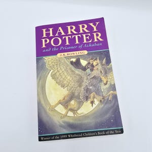 May include: A purple book cover with the title "Harry Potter and the Prisoner of Azkaban" by J.K. Rowling. The cover features an illustration of Harry Potter flying on a hippogriff against a night sky with a full moon.