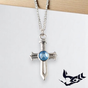May include: Silver-toned pendant necklace featuring a cross design with a blue gemstone center. The cross has a sword-like appearance and hangs from a delicate chain. The background is a soft white, with a wooden accent at the top.