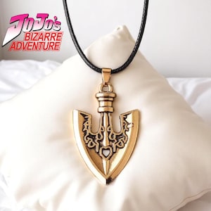 May include: Gold-colored pendant necklace with a black cord. The pendant is shaped like an arrow with intricate designs and a heart-shaped cutout. The necklace is displayed on a white pillow. The text "JoJo's Bizarre Adventure" is visible in the upper left corner.