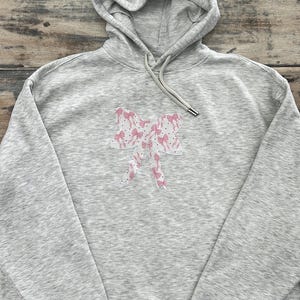 May include: A grey hooded sweatshirt with a pink bow design on the front. The bow is made of a patterned fabric with pink and white polka dots.