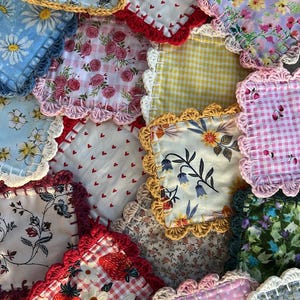 May include: A selection of handmade fabric coasters with diverse floral, fruit, and patterned designs. Each coaster has a crocheted edge in colours like yellow, red, and white. The coasters are square and made of cotton.