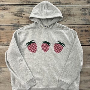 May include: A grey hooded sweatshirt with three red and white gingham strawberry appliques on the front.