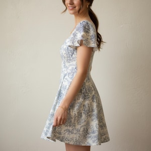 May include: A white and blue patterned dress with a flared skirt and short, ruffled sleeves. The dress features a detailed toile design, and a fitted bodice. The dress is knee-length and has a classic, feminine silhouette.