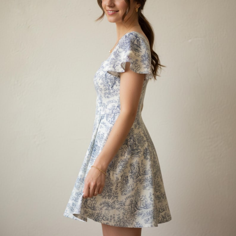May include: A white and blue patterned dress with a flared skirt and short, ruffled sleeves. The dress features a detailed toile design, and a fitted bodice. The dress is knee-length and has a classic, feminine silhouette.