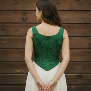 May include: A sleeveless emerald green bodice featuring a subtle tonal floral jacquard pattern, a scoop neckline, and a center back zipper closure, paired with a long, flowing cream-colored skirt that reaches toward the floor.