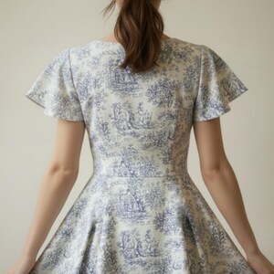 May include: A white and blue toile print dress with a flared skirt and flutter sleeves. The dress features a classic toile pattern with scenes of figures and landscapes. The dress is made of a lightweight fabric.