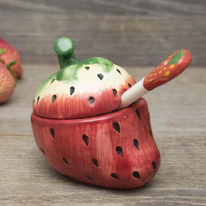 May include: A ceramic strawberry-shaped container with a lid and spoon. The container is red with black seed details, a green stem, and a cream-colored top. The spoon has a red strawberry design on the handle.