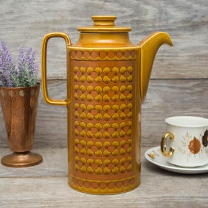 May include: A brown ceramic coffee pot with a geometric pattern of circles and flowers. The pot has a lid and a handle.