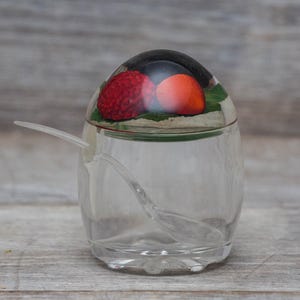 May include: A clear glass sugar dispenser with a red, orange, and black fruit design on the lid. The dispenser has a small spoon attached to the lid.