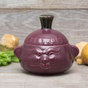 May include: A purple ceramic beetroot-shaped container with a lid. The container has a face with a grumpy expression and the word "BEETROOT" written on the lid.