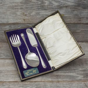 May include: A silver cutlery set in a black velvet-lined box. The set includes a fork, knife, and serving spoon. The box has a label that reads "Serve-All's Entree Fish Vegetable & Pastry Fruit"
