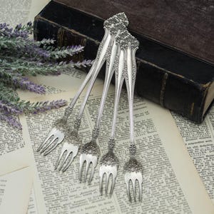 May include: A set of five silver-toned metal cocktail forks featuring ornate, embossed floral detailing on the handles and three tines. The forks are arranged in a fan-like pattern, showcasing the intricate scrollwork and polished metallic finish.
