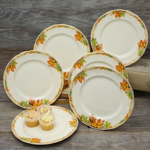 May include: A set of cream-coloured ceramic plates featuring a border of autumn leaves in orange, yellow, and green. The plates are arranged on a wooden surface, with a small plate holding cupcakes.