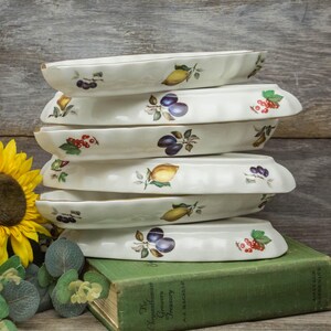 May include: A stack of six white oval serving dishes. Each dish is decorated with a different fruit design, including lemons, plums, and red currants. The rims are trimmed with gold. The dishes are stacked on a green book.