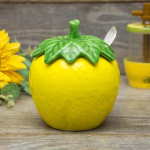 May include: A ceramic lemon-shaped sugar bowl with a green leaf-shaped lid and a small spoon. The bowl is bright yellow, and the lid is green. The sugar bowl is on a wooden surface.