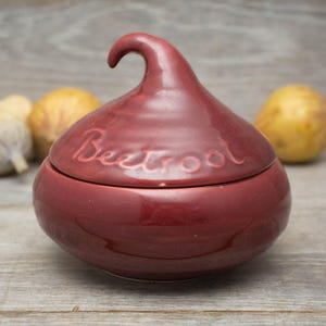 May include: A burgundy ceramic beetroot-shaped container with a lid. The lid has a curved handle and the word "Beetroot" is written on the top.