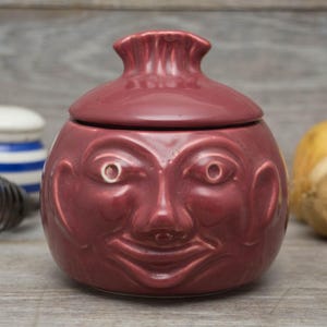 May include: A burgundy ceramic jar with a lid shaped like a face. The face has a wide smile, large eyes, and a prominent nose.