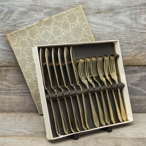 May include: A set of gold-coloured cutlery, including knives and forks, presented in a white box with a black interior. The box is open, revealing the utensils. The box is next to a decorative, patterned case.
