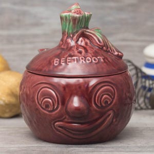 May include: A red ceramic beet-shaped sugar bowl with a green leafy top and a smiling face. The lid has the word "BEETROOT" embossed on it.