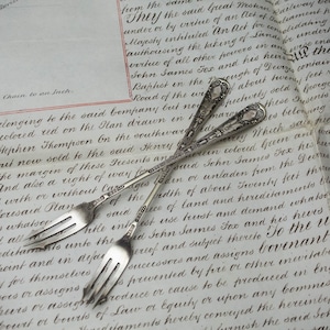 May include: Two silver-toned metal forks with three tines each, featuring intricate embossed floral and scrollwork patterns along the elongated handles. The forks are positioned in a crossed arrangement, showcasing detailed craftsmanship on the stems.