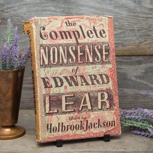 May include: A vintage copy of "The Complete Nonsense of Edward Lear" edited by Holbrook Jackson. The book has a worn, aged cover with a red and cream color scheme and decorative illustrations. The book is propped up on a small black stand.