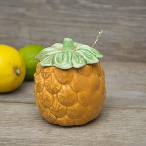 May include: A ceramic pineapple-shaped sugar bowl with a green leaf lid and a small spoon. The bowl is a light brown colour and has a textured surface.