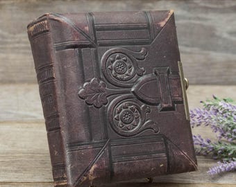 Puede incluir: Un libro antiguo, encuadernado en cuero marrón oscuro con diseños florales y geométricos en relieve. El libro tiene un cierre metálico y está ligeramente inclinado, mostrando su detallada cubierta. Los bordes muestran signos de desgaste, lo que añade encanto antiguo.