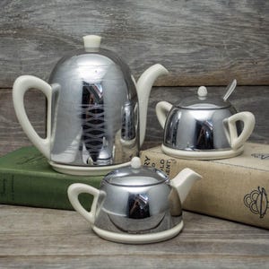 May include: A chrome tea set comprising a teapot, sugar bowl, and creamer, each with white handles and bases. The items are displayed on old books against a backdrop of aged wooden planks. The teapot has a decorative wave pattern.
