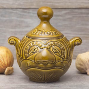 May include: An olive green ceramic onion storage pot. The lidded container has a face design and the word "Onions" on the lid. It features two side handles and a round knob. A decorative kitchen item.
