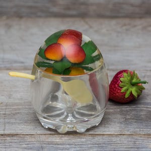 May include: A clear glass sugar dispenser with a lid featuring a pressed image of red and green strawberries. The dispenser has a small yellow spoon inside and sits on a wooden surface.