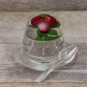 May include: A clear glass dome with a lid and a small spoon. Inside the dome are three red berries and green leaves.