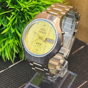 May include: A gold-toned Orient watch with a yellow face, date display, and a stainless steel band. The watch is displayed on a clear stand, with a green plant in the background. The watch face has the word "Orient" and three stars.