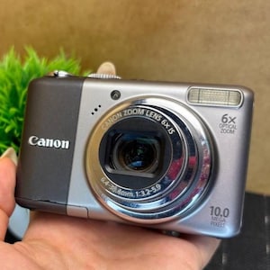 May include: A silver and black Canon digital camera with a zoom lens. The camera has a 10.0 megapixel resolution and 6x optical zoom. The Canon logo is visible on the front of the camera.