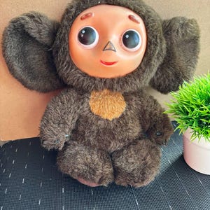 以下が含まれることがあります： 茶色の毛むくじゃらの体、大きな耳、大きな目と小さな鼻を持つ丸い顔が特徴のぬいぐるみ。顔は明るい桃色で、赤い笑顔です。おもちゃの胸には、小さくて丸いオレンジ色のパッチが付いています。
