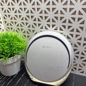 Könnte beinhalten: Ein silberner, tragbarer Sony Walkman CD-Player mit kreisförmigem Design. Das Gerät verfügt über ein markantes Sony-Logo auf dem oberen Deckel, ein blaues Walkman-Emblem und mehrere Bedientasten am unteren Rand, einschließlich einer Öffnungstaste und Wiedergabesteuerungen.