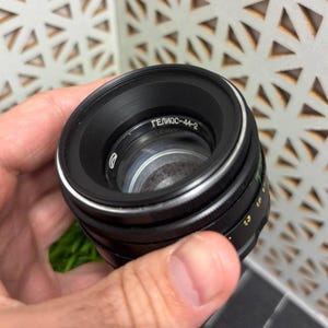 Peut inclure: Un objectif d'appareil photo noir avec le texte "ГЕЛИОС-44-2" est tenu dans une main. L'objectif a un anneau argent&eacute; et des marquages verts et blancs. L'objectif est con&ccedil;u pour la photographie.