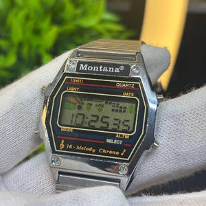 以下が含まれることがあります： シルバーカラーのMontanaクォーツデジタル腕時計。金属製のリンクバンド付き。長方形の文字盤には黒いディスプレイがあり、Montana、Quartz、16-Melody Chrono、Light、Date、Mode、Select、Al.Tmの文字が表示され、時刻は10時25分35秒を示しています。