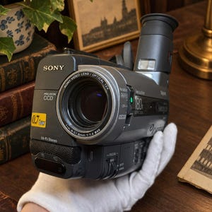 May include: A dark gray Sony Handycam camcorder featuring a 12x optical zoom lens, a Precision CCD sensor, and a 180x digital zoom capability. The device includes a 14-Fx Stereo microphone, a 0.7 Lux rating, and a top-mounted electronic viewfinder.