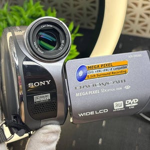 以下が含まれることがあります： カールツァイス バリオ・テッサーレンズを搭載したグレーのソニー製ハンディカムデジタルビデオカメラ。フリップアウト画面には、Mega Pixel、12x Optical Zoom、Wide LCD、DVD R/RW互換性などのテキストが表示されています。Dolby Digital 5.1 Creatorを搭載しています。