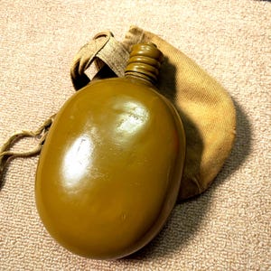 May include: A green metal canteen with a brown canvas carrying case. The canteen has a screw-on cap and a small carrying strap.