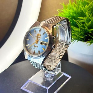 May include: A vintage Orient watch with a silver-tone case and mesh band. The watch face is light blue with gold-tone hands and markers. The day and date are displayed at the 3 o'clock position. The watch is displayed on a clear acrylic stand.