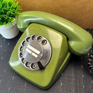 May include: An olive green rotary telephone featuring a circular dial with black numbers from zero to nine. The handset rests on top of the base, which has a central circular plate with two horizontal white lines. The device has a glossy finish.