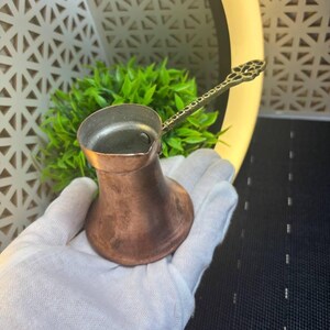 May include: A small copper coffee pot with a wide base, a narrow neck, and a long, slender handle featuring an intricate, perforated metal design. The exterior of the pot has a warm, metallic finish with visible signs of natural oxidation.