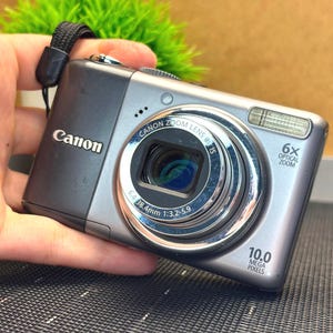 以下が含まれることがあります： 黒いリストストラップが付いたシルバーのキヤノンデジタルカメラ。カメラは6倍光学ズームレンズと10.0メガピクセルを備えています。キヤノンのロゴがカメラの前面に表示されています。