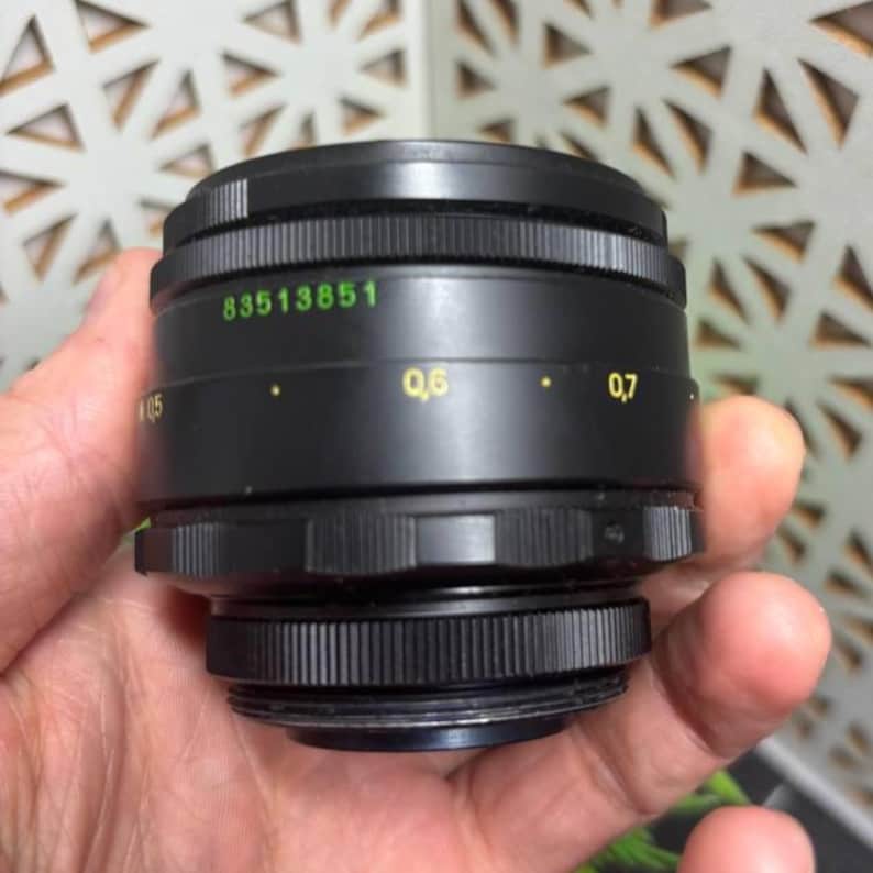 Peut inclure: Un objectif d'appareil photo noir avec des bords stri&eacute;s et une base filet&eacute;e. L'objectif porte des marquages verts et jaunes, dont le num&eacute;ro "83513851". L'objectif est tenu dans une main.