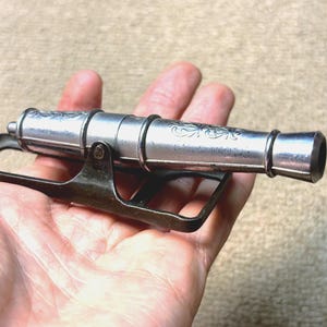 May include: A miniature silver cannon with decorative floral engravings on the barrel, mounted on a dark metal carriage. The cannon is held in a hand, showcasing its small size and intricate details. A vintage collectible.
