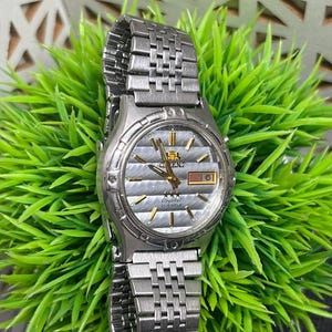 May include: Silver-toned Orient watch with a textured silver face, gold accents, and a day-date display. The watch has a stainless steel band and is set against a backdrop of green foliage.
