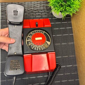 May include: Vintage red and black rotary dial telephone with a black handset. The phone has a circular dial with numbers and a red center. The handset is held up, showing the speaker and microphone. A green plant in a white pot is in the background.