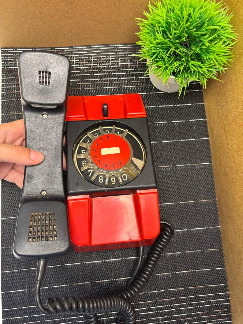 May include: Vintage red and black rotary dial telephone with a black handset. The phone has a circular dial with numbers and a red center. The handset is held up, showing the speaker and microphone. A green plant in a white pot is in the background.