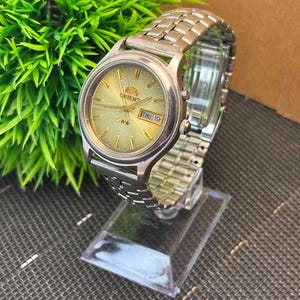 May include: A silver-toned Orient wristwatch with a gold-colored face and a stainless steel band. The watch face displays the Orient logo, two stars, and the day of the week. The watch is displayed on a clear stand.